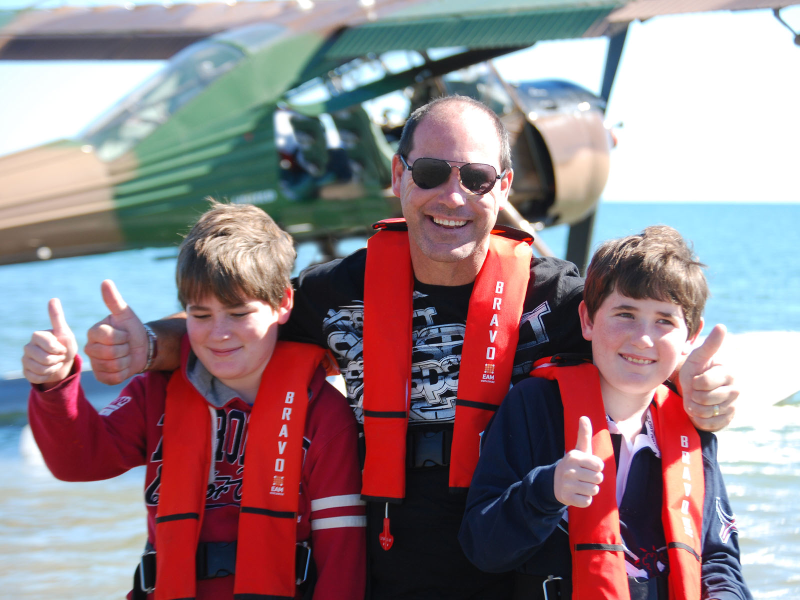 Two boys with their dad, wearing red life vests and standing in the water in front of seaplane.